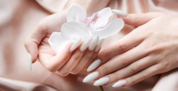 Beautiful hands of a young woman with white manicure on nails. Beige silk background and orchid flowers